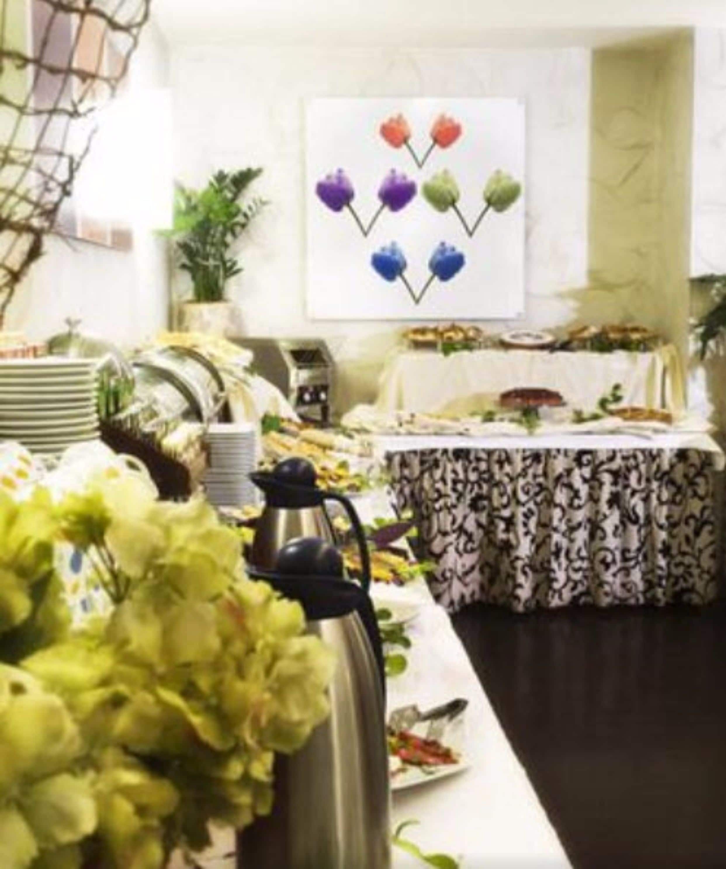 A dining area with a long table set for a meal, a decorative wall piece above, and a potted plant to the side.