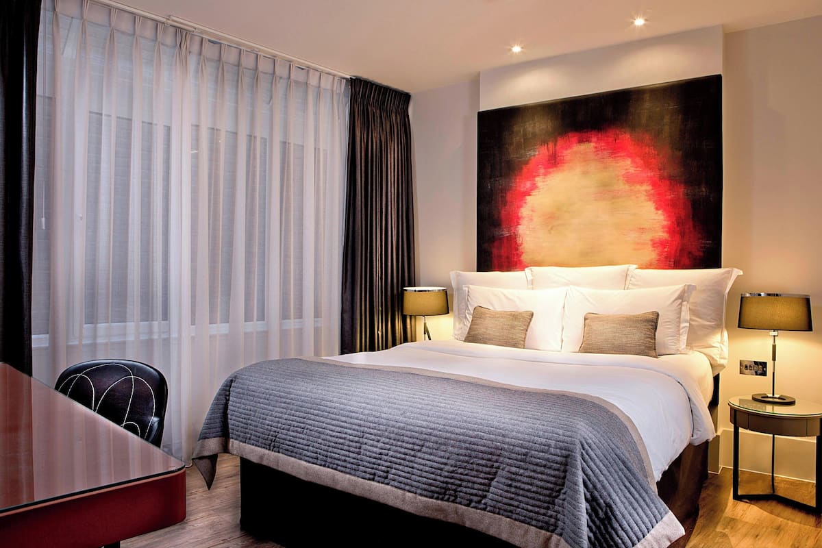 Een hotelkamer met een groot bed, een bureau en een stoel. Er is een raam met gordijnen en een schilderij aan de muur.