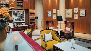 Un vestíbulo amplio con un sofá rojo, sillones amarillos y una mesa de centro de mármol. Se observan estanterías y cuadros enmarcados en las paredes.
