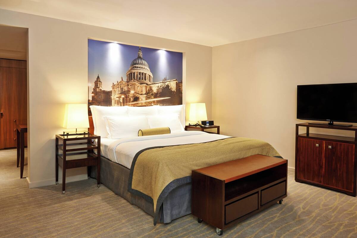Een hotelkamer met een groot bed, een flatscreen-televisie, een houten nachtkastje, een lamp en een poster van een gebouw aan de muur.