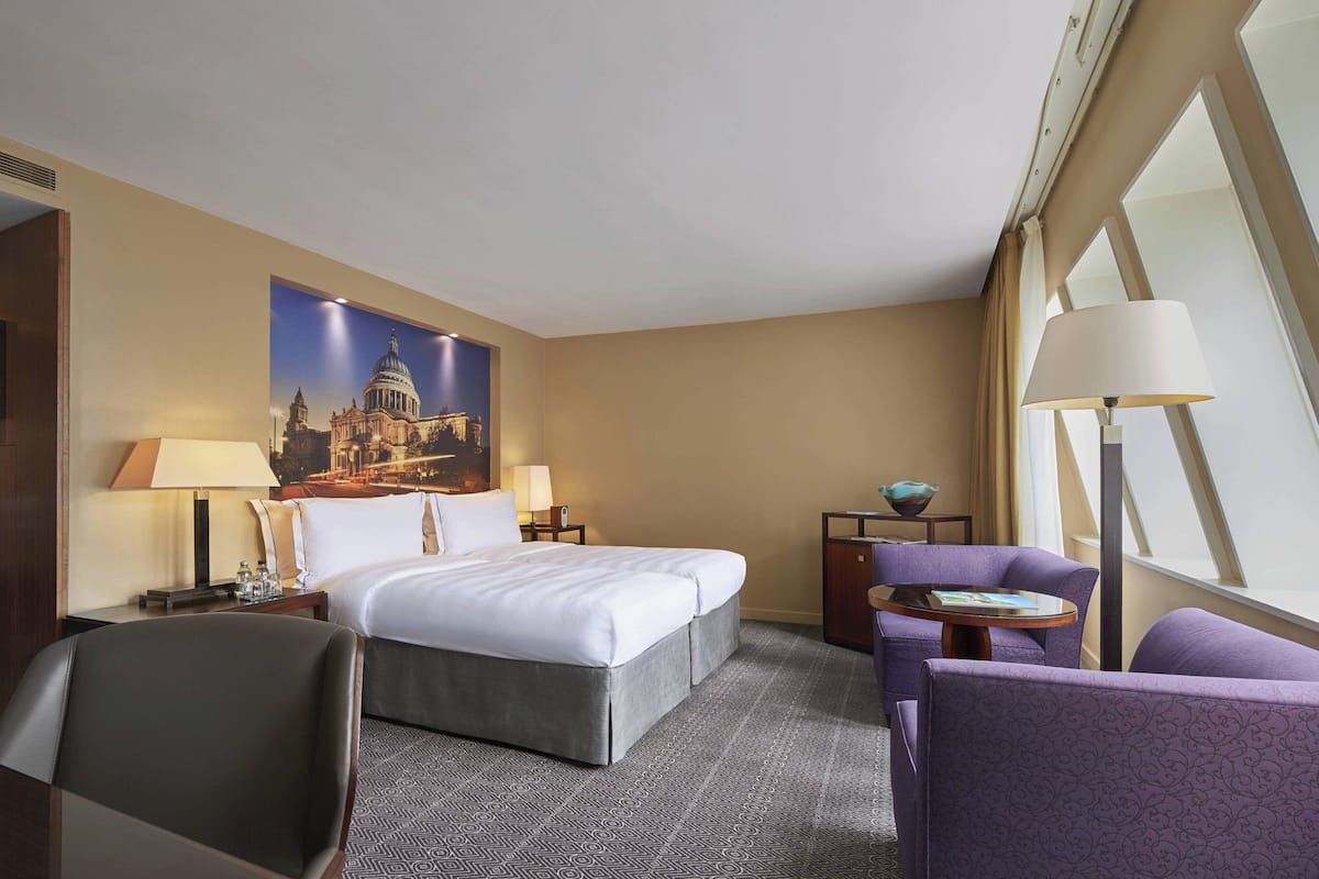 Hotelkamer met een groot bed, twee nachtkastjes met lampen, een paarse fauteuil, een klein rond tafeltje en een groot raam met gordijnen.