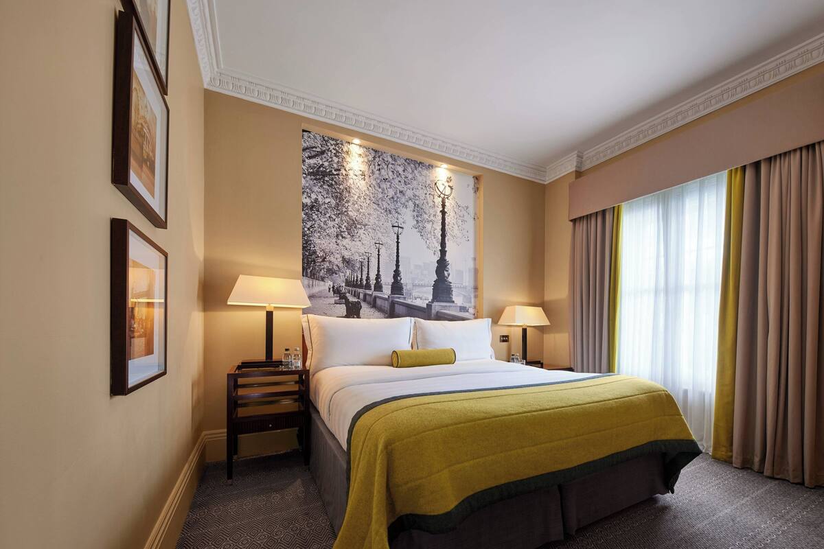 Een hotelkamer met een groot bed, een nachtkastje met een lamp, een aan de muur hangend schilderij en een raam met gordijnen.