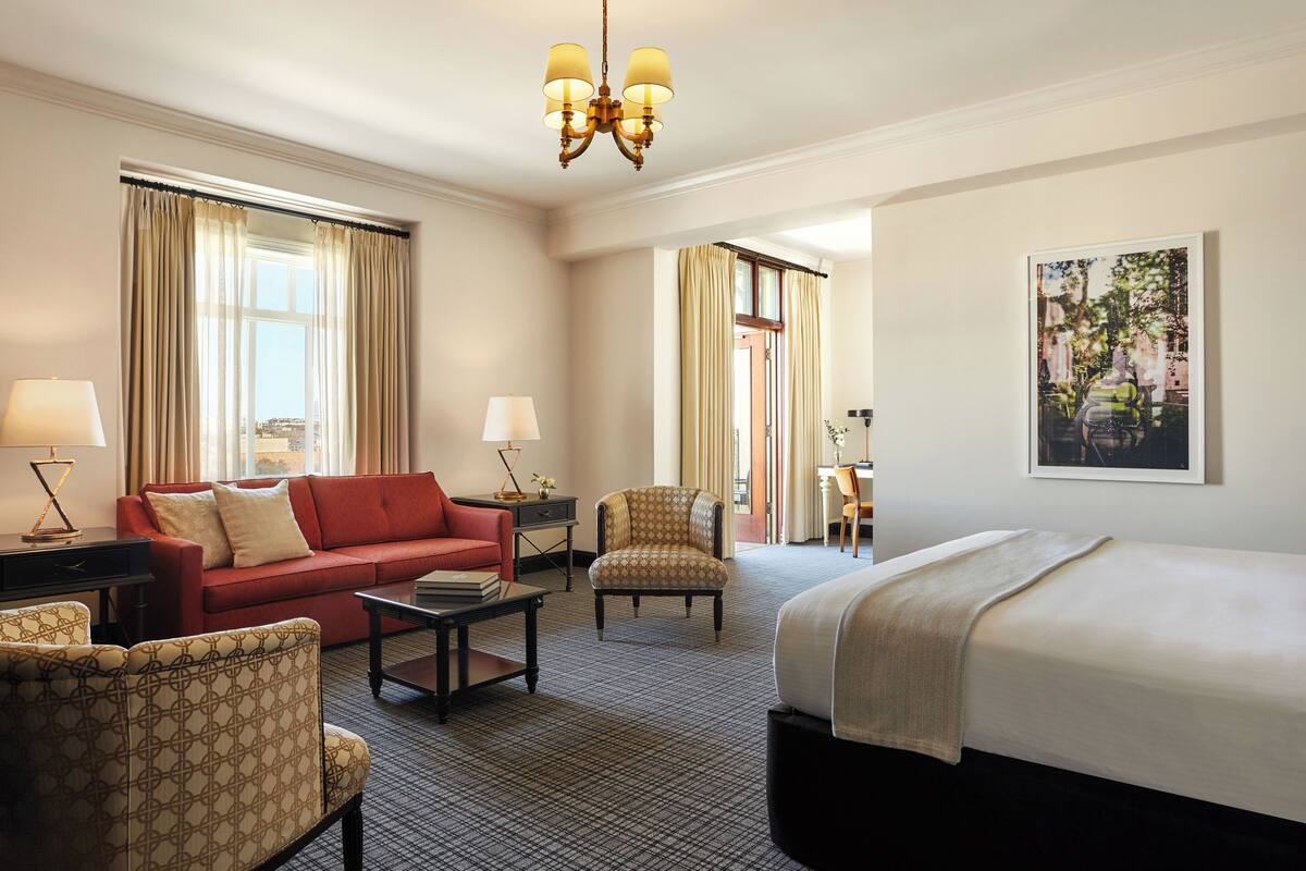 A hotel room with a bed, a red sofa, a patterned armchair, a small table, and a desk with a lamp.