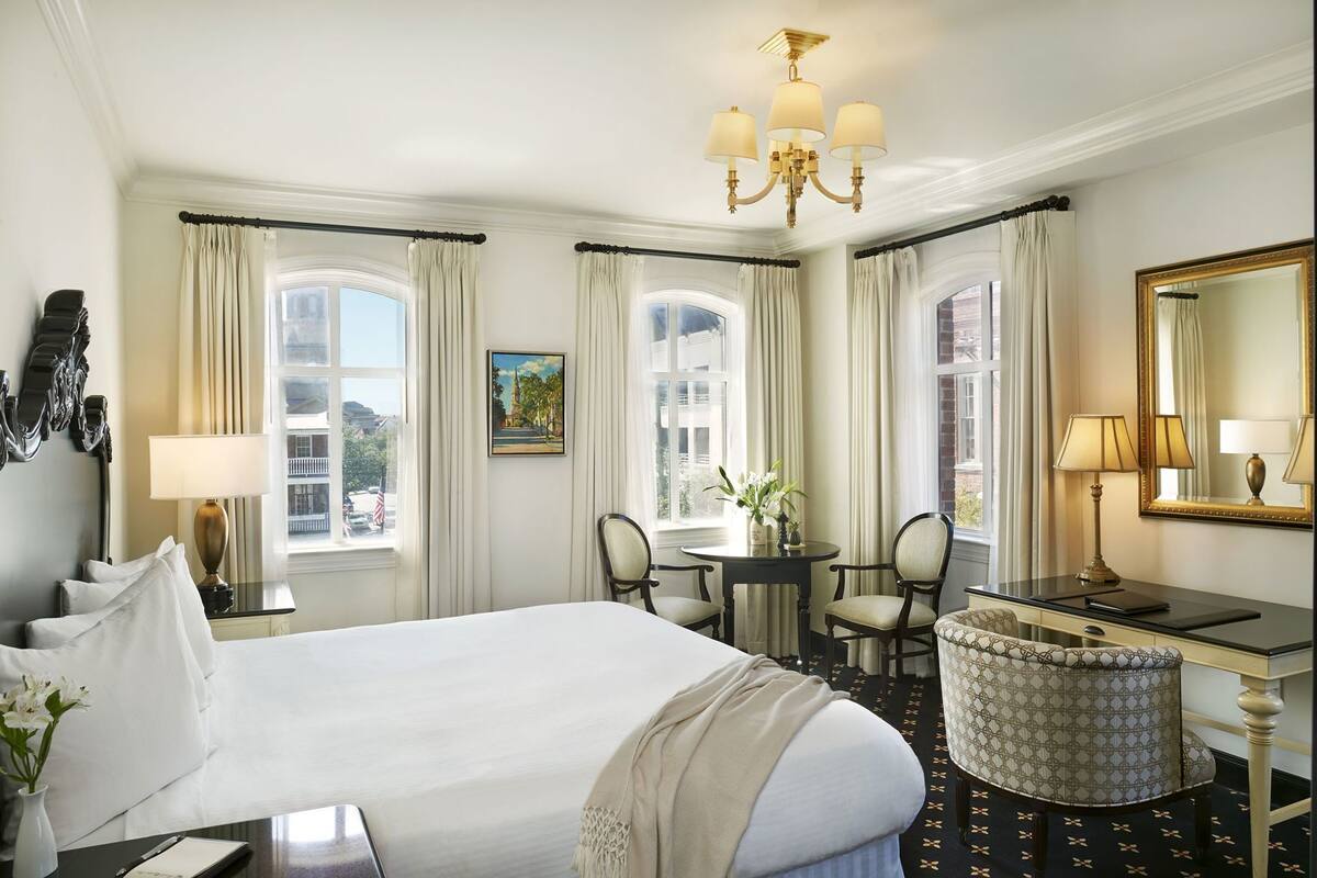 A hotel room with a large bed, two chairs, a desk, a mirror, and a chandelier.