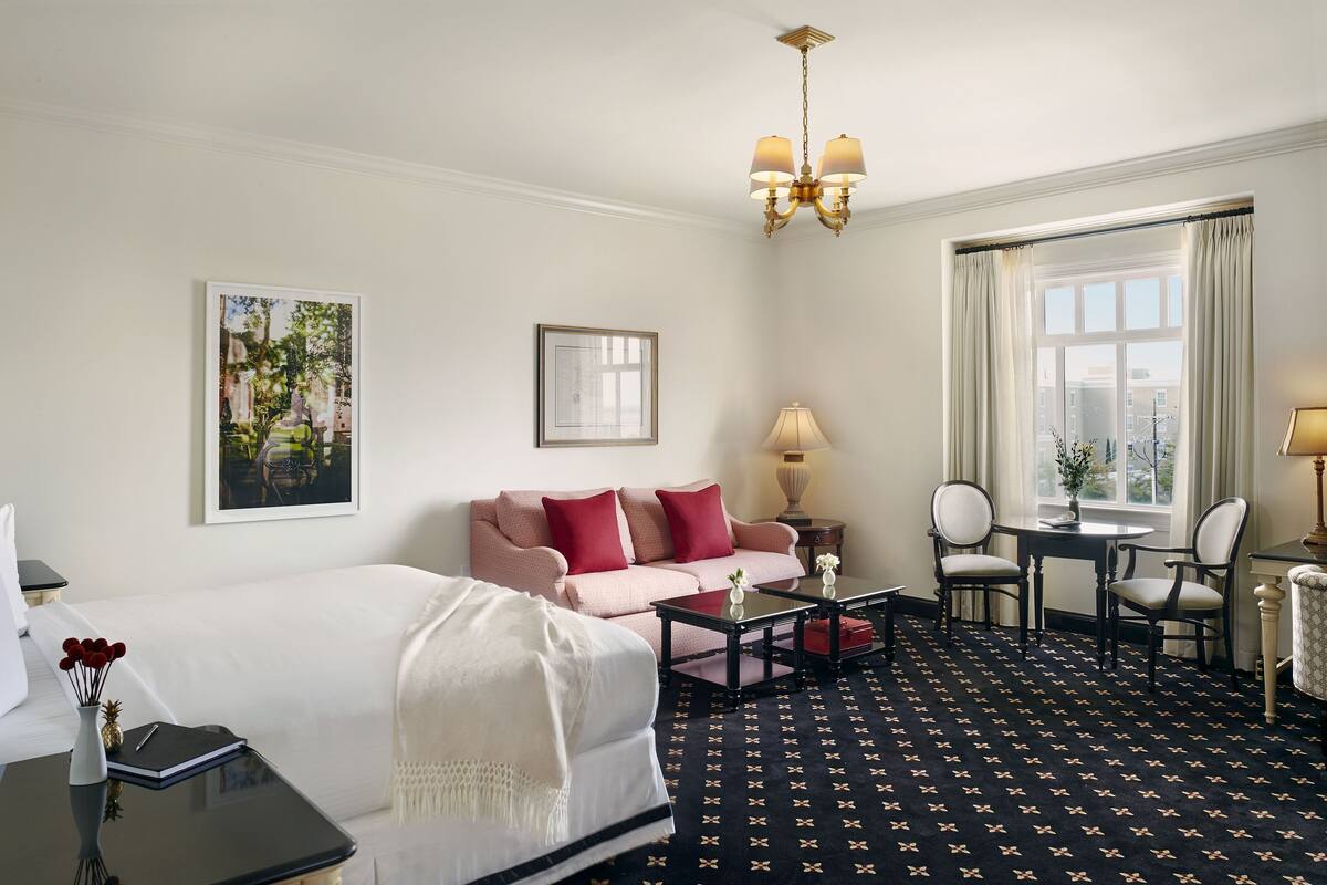 A hotel room with a bed, a sofa, a dining table with chairs, a chandelier, and a large window with curtains.