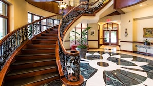 Una gran escalera con detalles de madera y barandillas de metal ornamentadas, que conduce a un vestíbulo con piso de mármol y plantas en macetas.