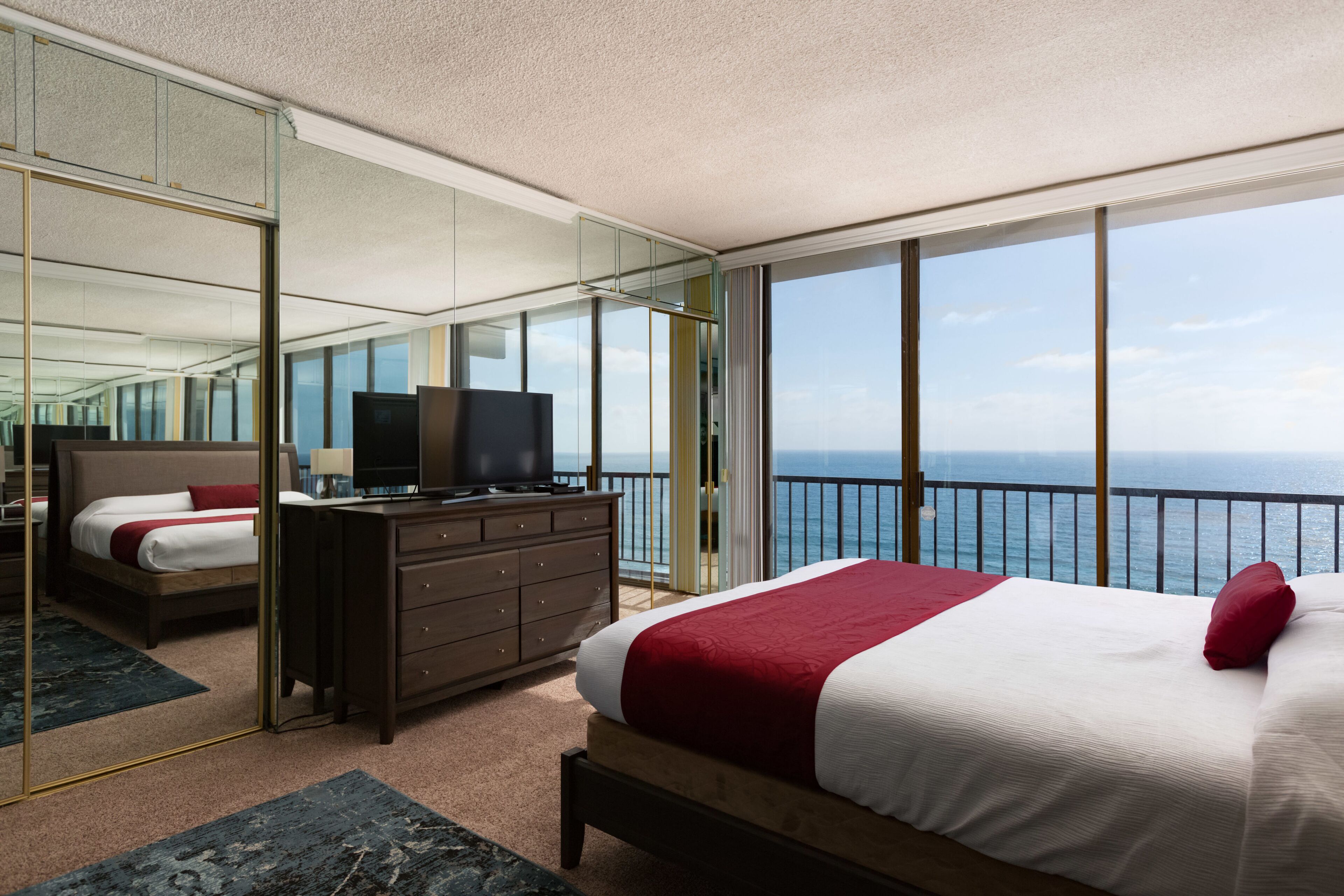 A modern hotel room with a large bed, a dresser, a TV, and a balcony with a view of the sea.