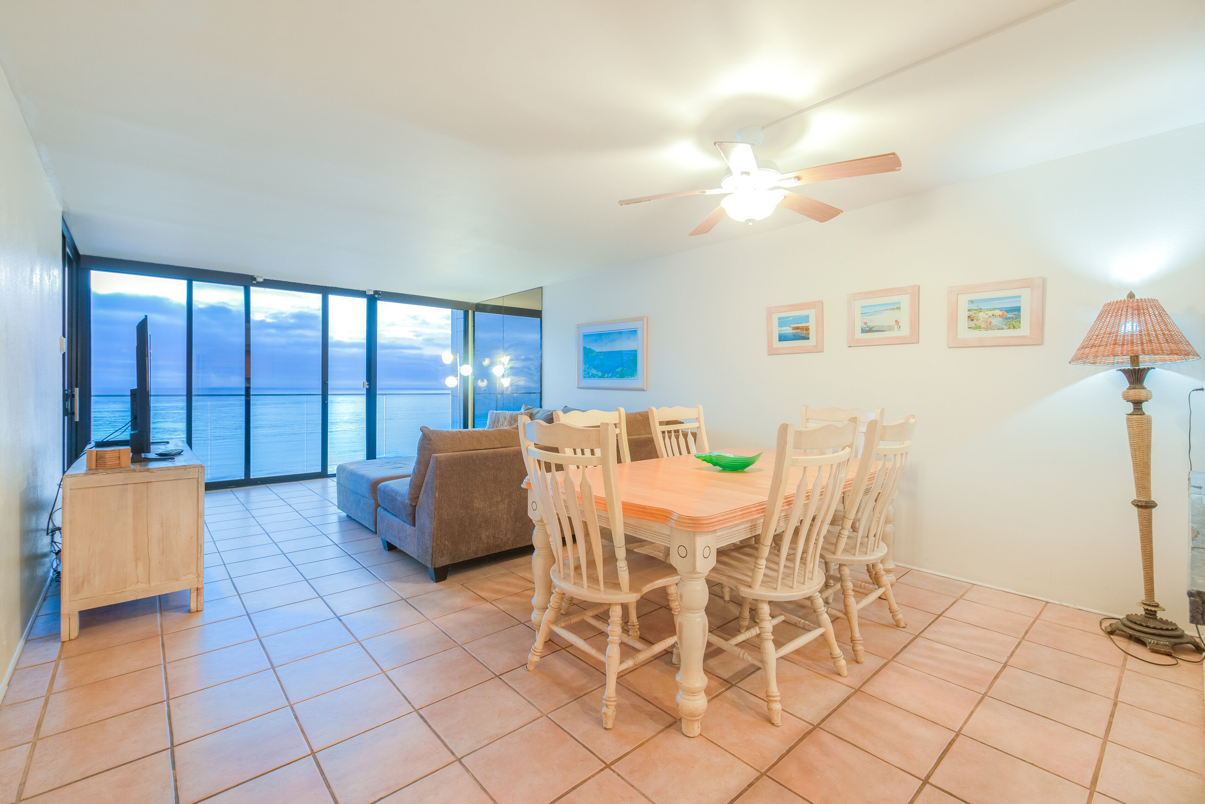 A dining area with a table and chairs, a ceiling fan, and a view of the ocean through large windows.