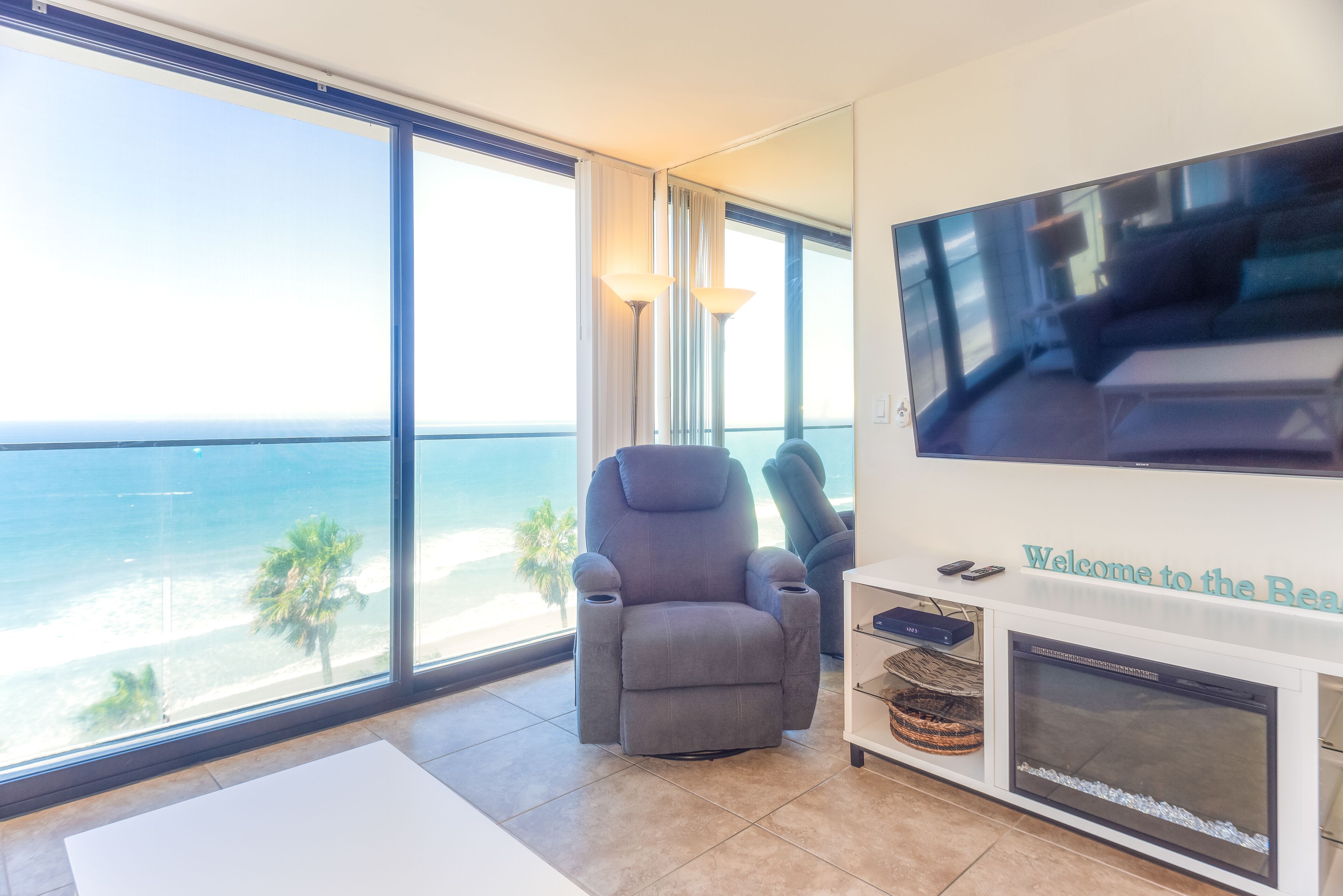 A living room with a large window overlooking the beach, a grey armchair, a flat-screen TV, and a fireplace.