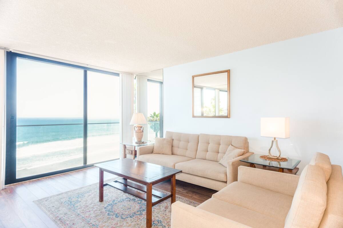 Amplia sala de estar con un gran ventanal con vista a la playa, un sofá color beige, una mesa de centro de madera y una mesita auxiliar con lámpara.