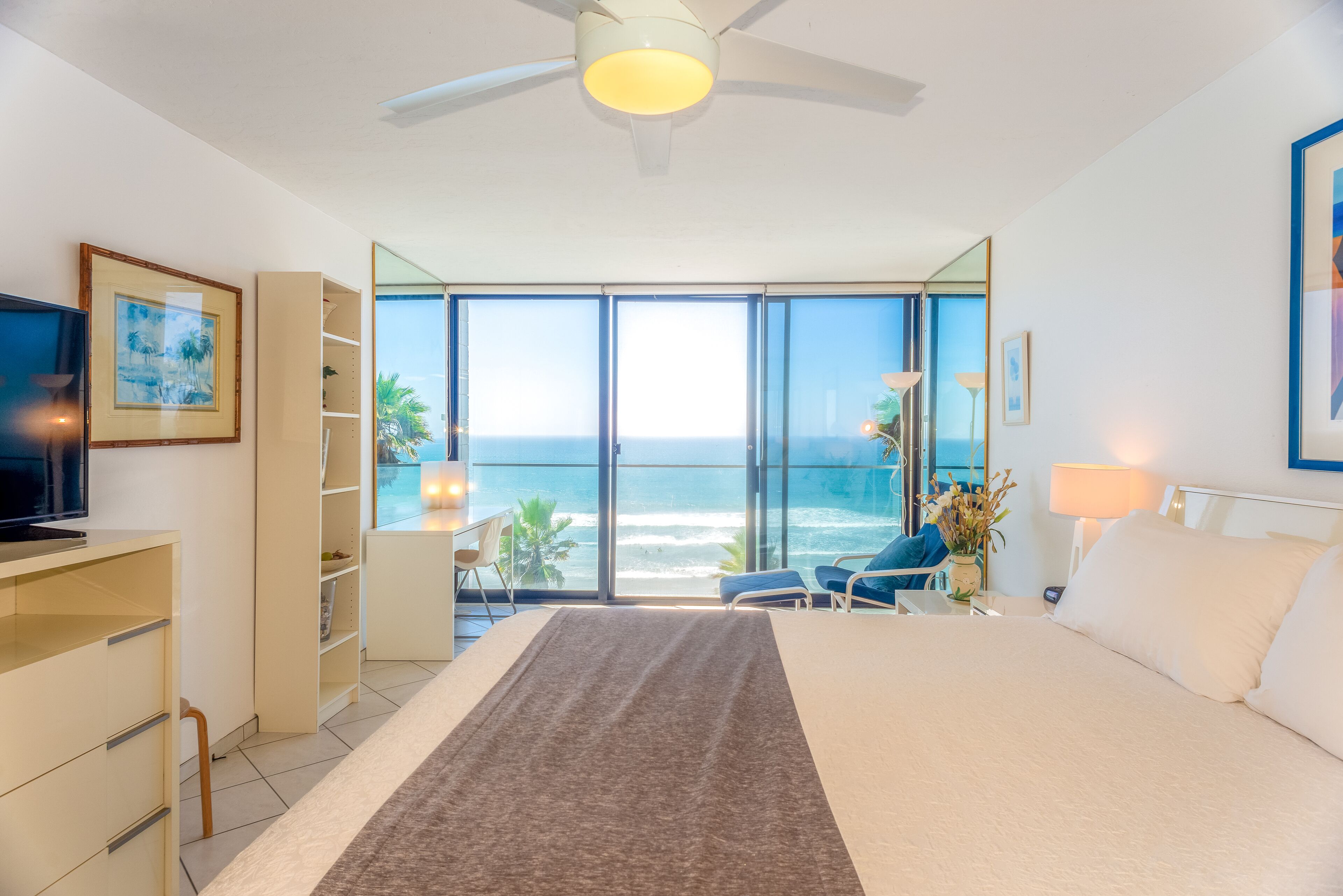 A bedroom with a large bed, a desk, a TV, and a view of the ocean through large windows.