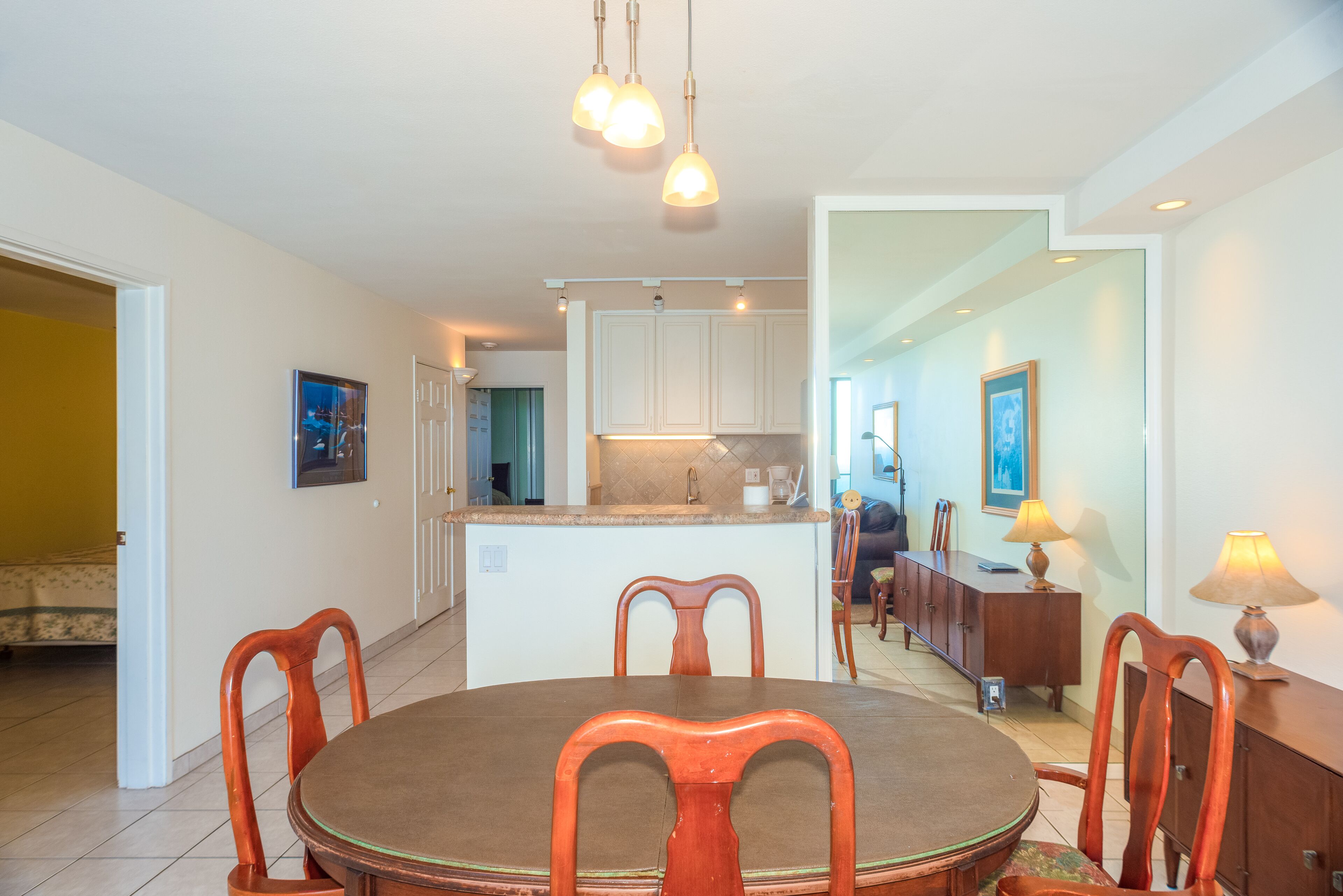 A dining area with a round table, wooden chairs, a kitchen in the background, and pendant lighting.