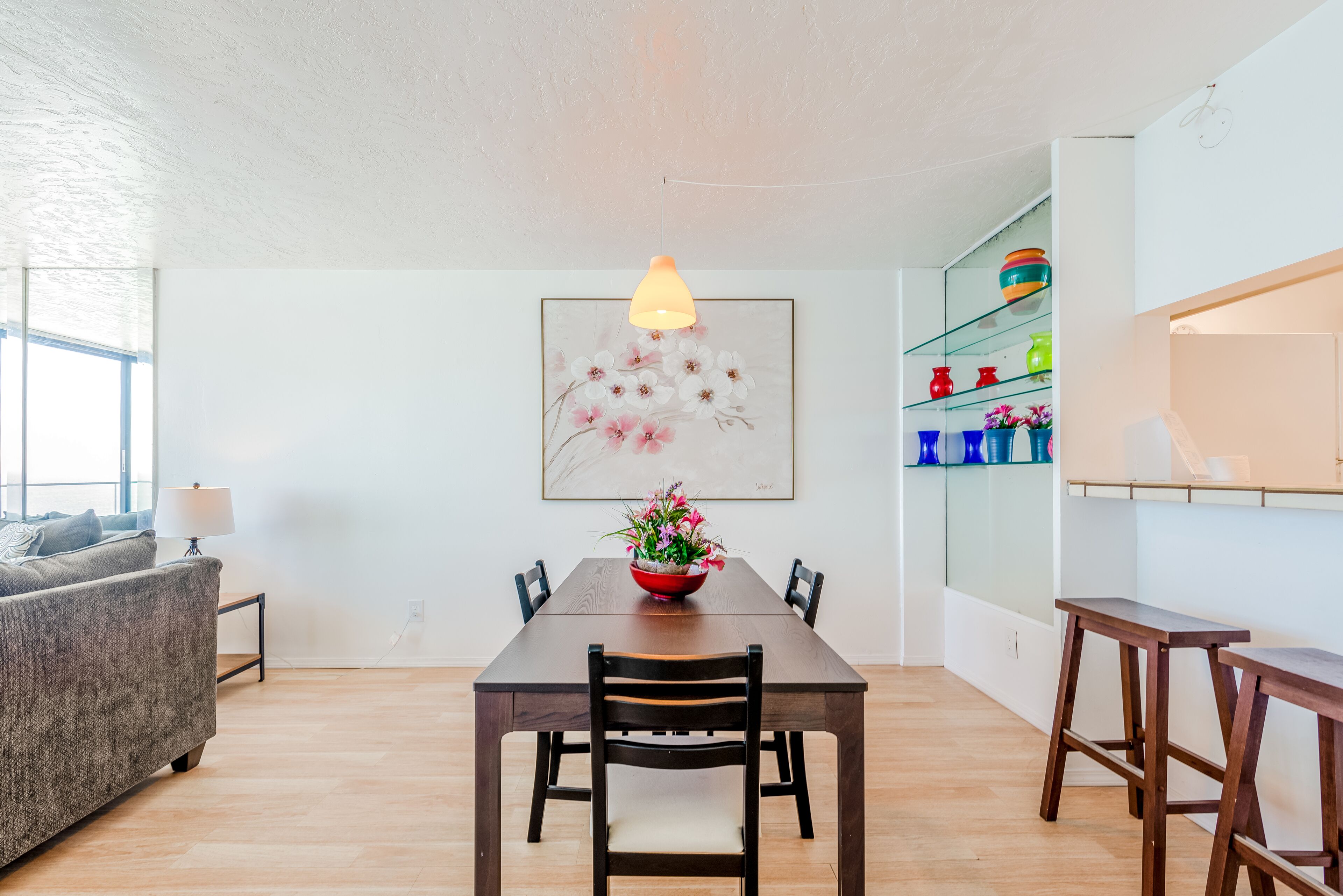 A dining area with a wooden table, chairs, a hanging pendant light, a floral painting, and a shelf with colorful items.