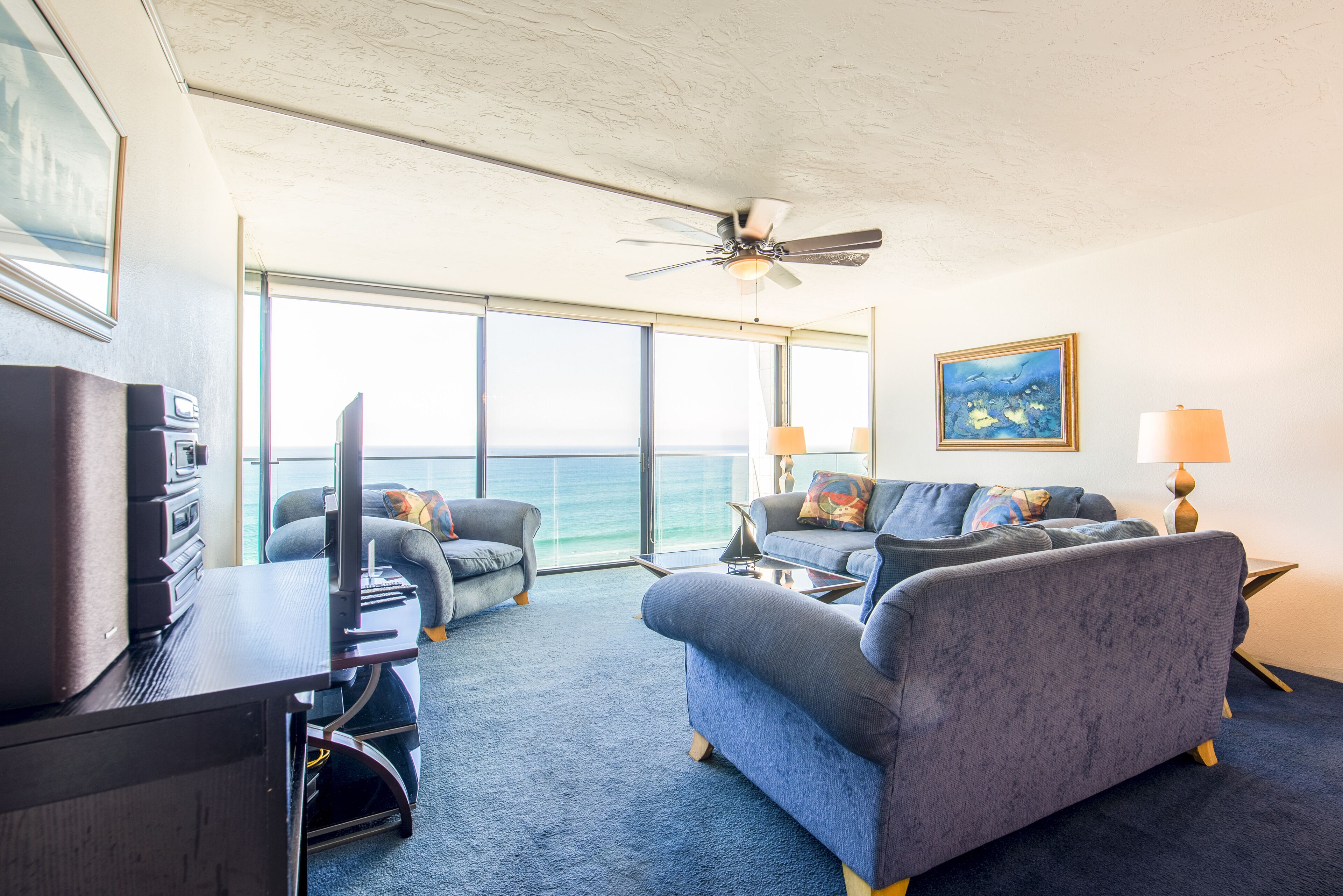 A room with a large window, a ceiling fan, a desk with a computer, two sofas, and a painting on the wall.