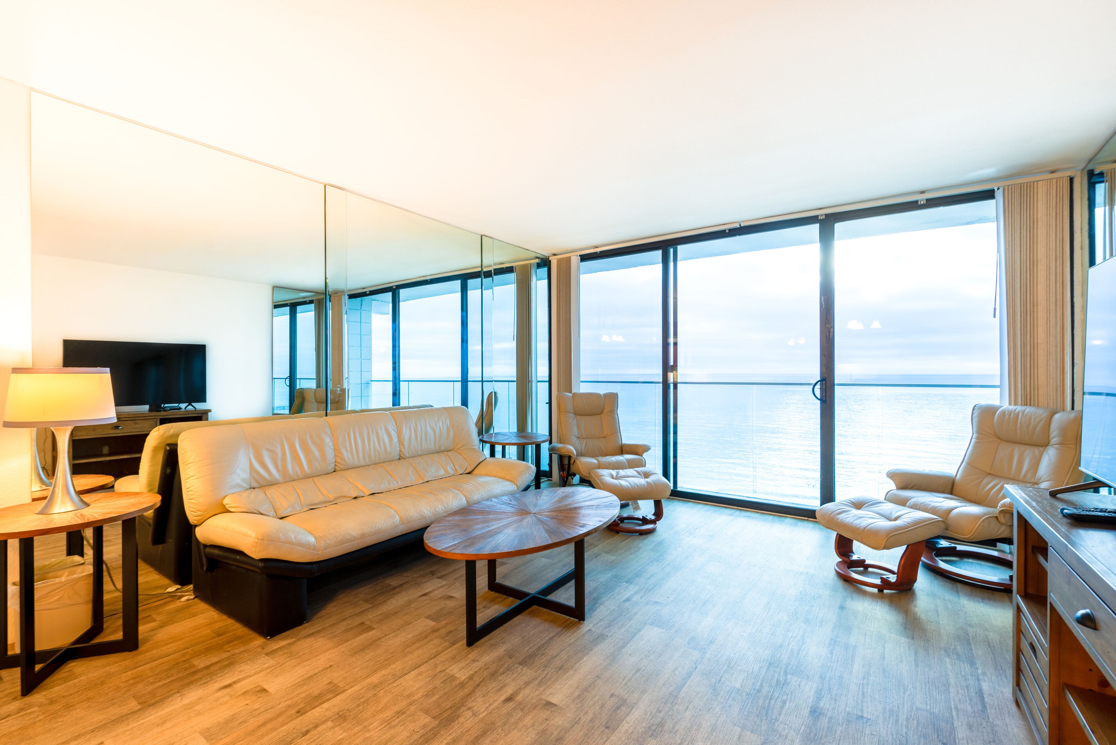 Une chambre d’hôtel moderne dotée d’une grande fenêtre donnant sur la mer, d’un canapé, d’un fauteuil, d’une petite table et d’un téléviseur.