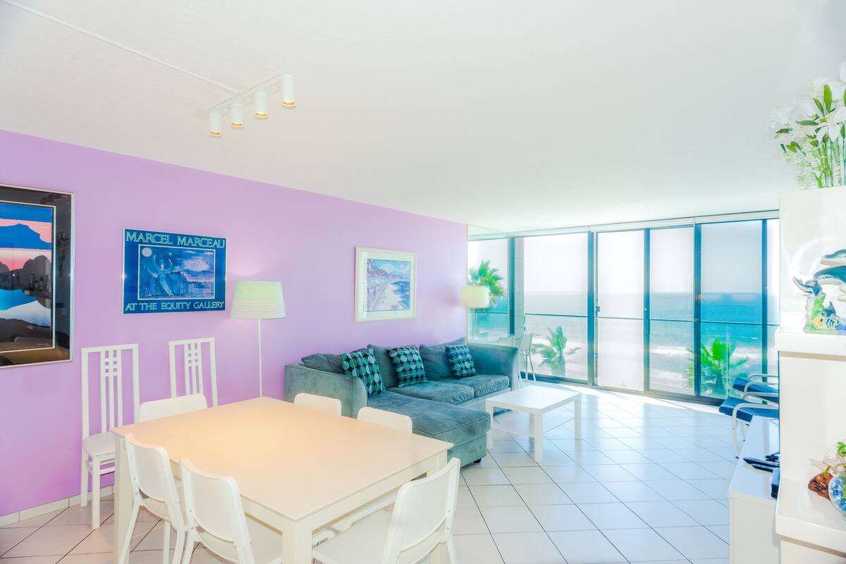 Un salón luminoso con un ventanal grande con vistas a la playa, un sofá, una mesa de comedor y algunas sillas.