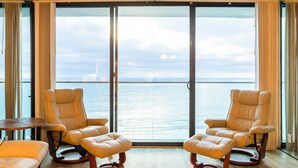 Ein Zimmer mit zwei Ledersesseln, einem kleinen Tisch, großen Fenstern mit Vorhängen und Blick auf das Meer.