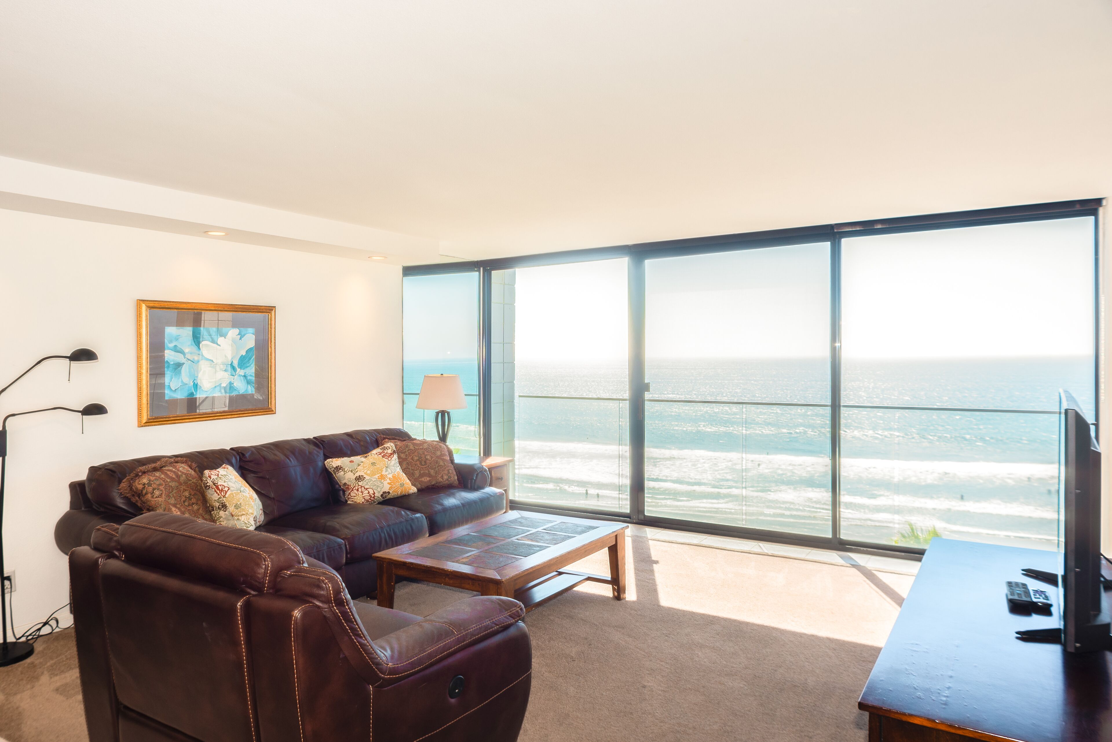 A modern living room with a leather sofa, a coffee table, and a TV on a wooden desk. Large windows offer a view of the ocean.