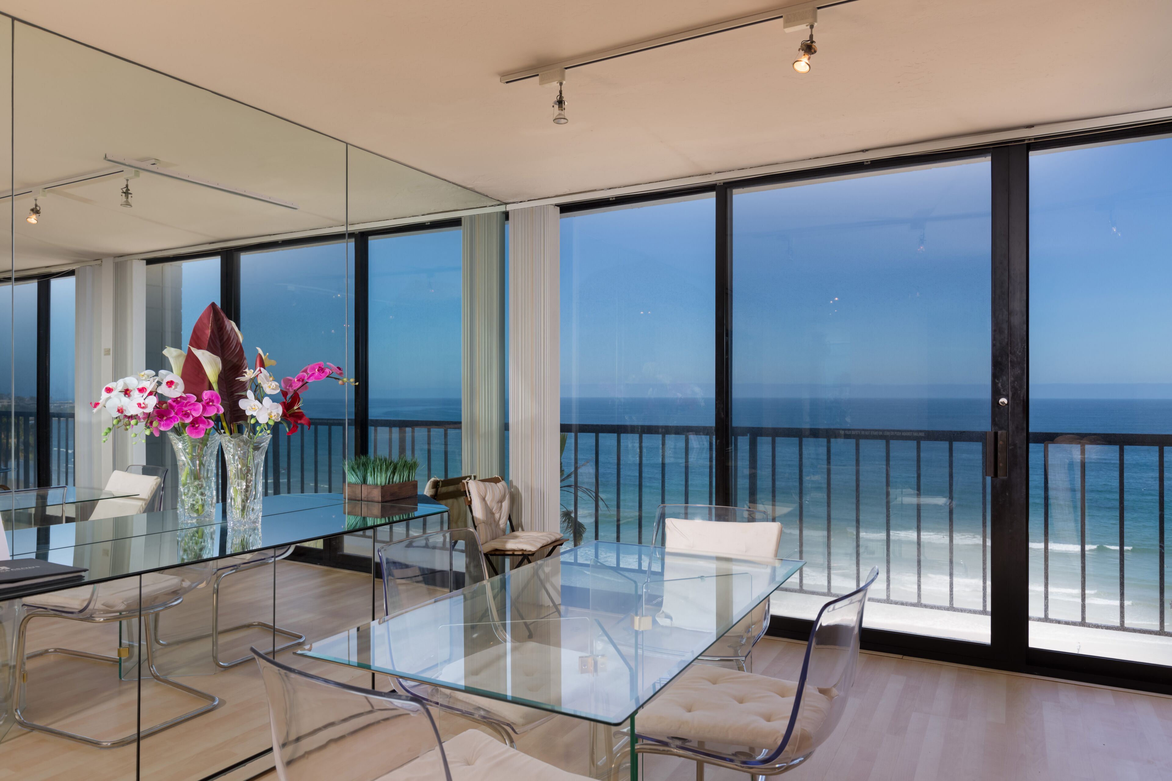 A modern dining area with a glass table and chairs, large windows with a view of the ocean, and a balcony with a railing.