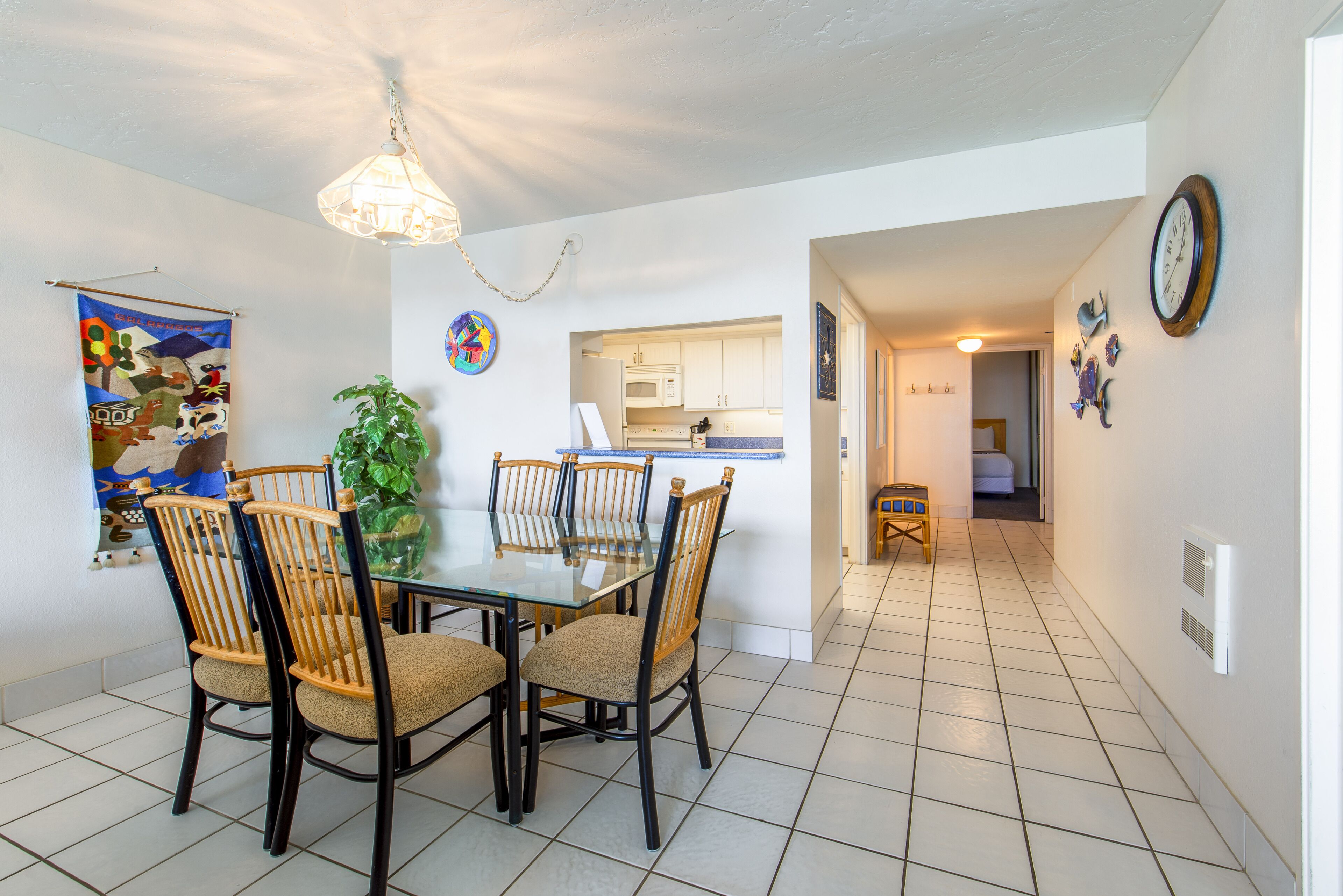 A dining area with a glass table and wooden chairs, a kitchen in the background, and a hallway leading to a bedroom.