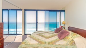 Ein Schlafzimmer mit einem großen Bett, einem Schminktisch und Meerblick durch raumhohe Fenster.