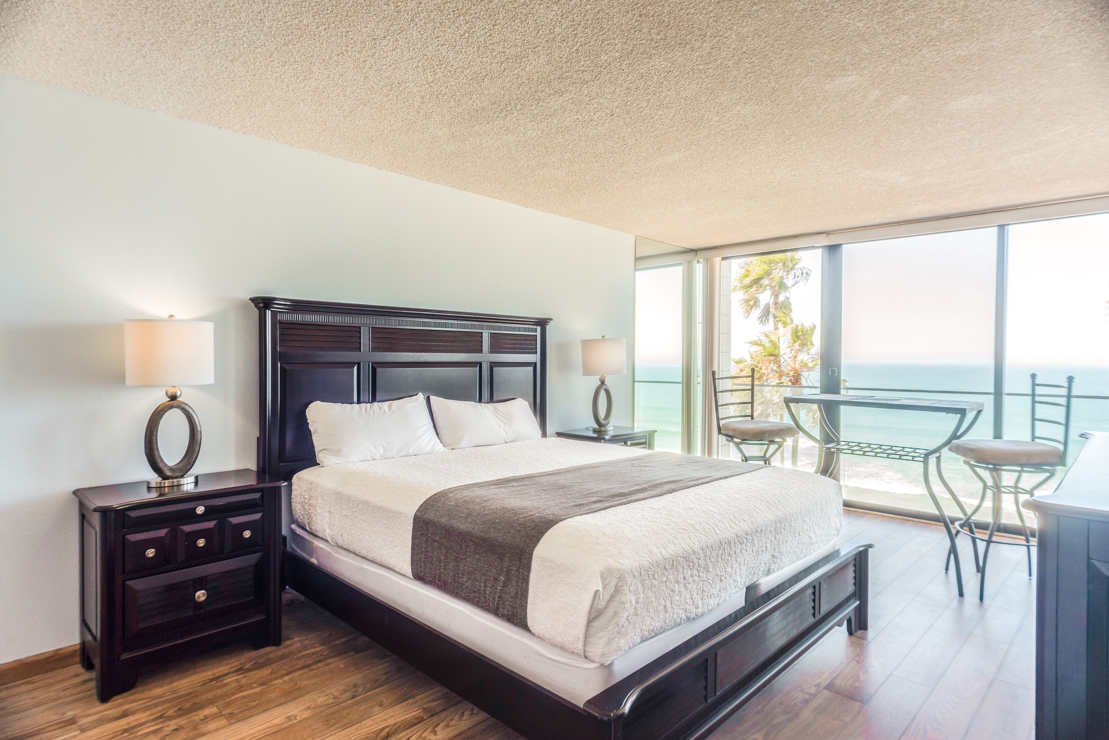 A bedroom with a large bed, two bedside tables, a nightstand, and a balcony with a table and chairs.