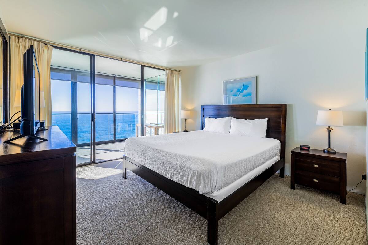 Habitación de hotel con una cama grande, un escritorio y vista al océano a través de ventanas de piso a techo.