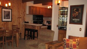 A cozy living room with a brown sofa, a wooden coffee table, and a patterned throw pillow. A wooden dining table with chairs is in the background, and a kitchen area with wooden cabinets and a microwave is visible. - Val d'Isere by Mammoth Reservation Bureau (Mammoth Lakes)