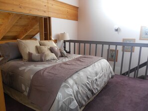 Et soveværelse med træloft, en seng med mange puder, et natbord med lampe og udsigt til en balkon med gelænder.