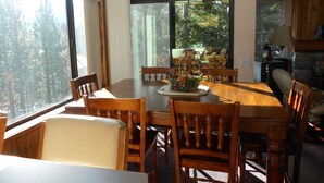 A dining area with a wooden table and chairs, large windows with a view of trees, and a stone fireplace. - Val d'Isere by Mammoth Reservation Bureau (Mammoth Lakes)