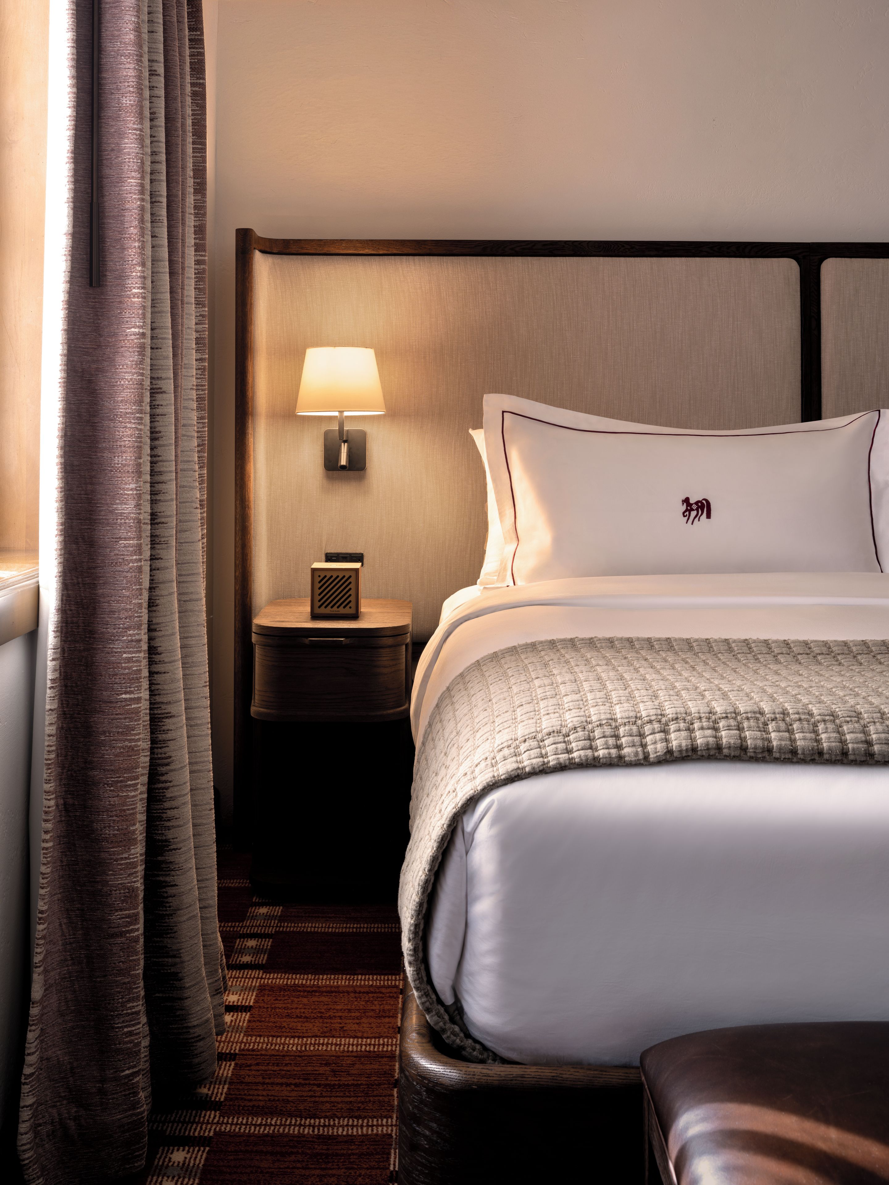 A hotel room with a bed, a nightstand, a lamp, and a window with curtains.