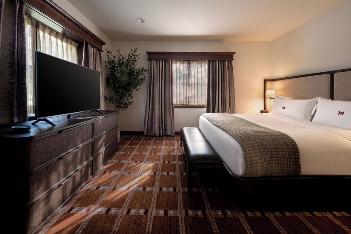A hotel room with a large flat-screen TV, a bed with white linens, a bench, a plant, and a window with curtains.