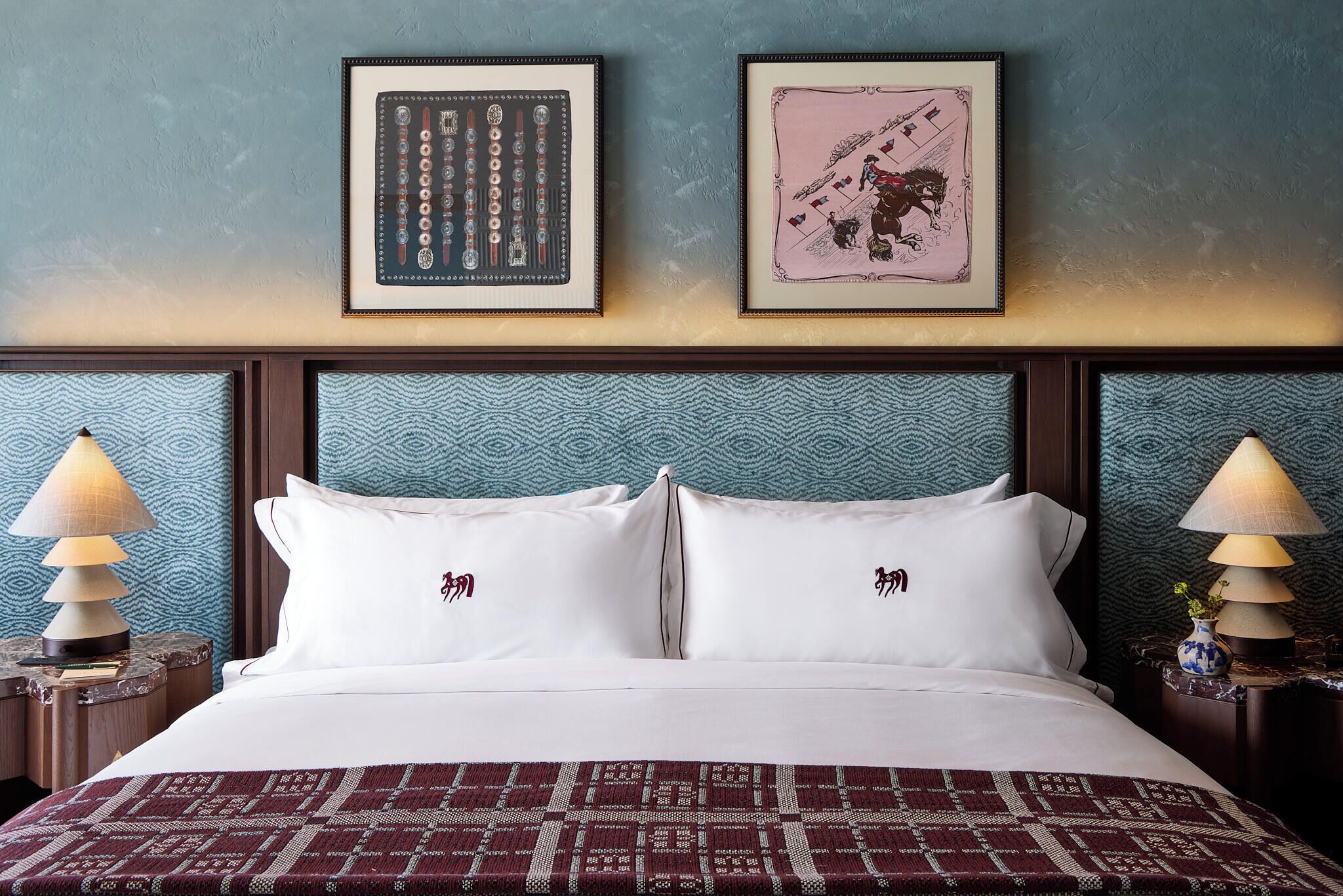 Quarto de hotel com uma cama, duas molduras na parede e duas luminárias de cabeceira.