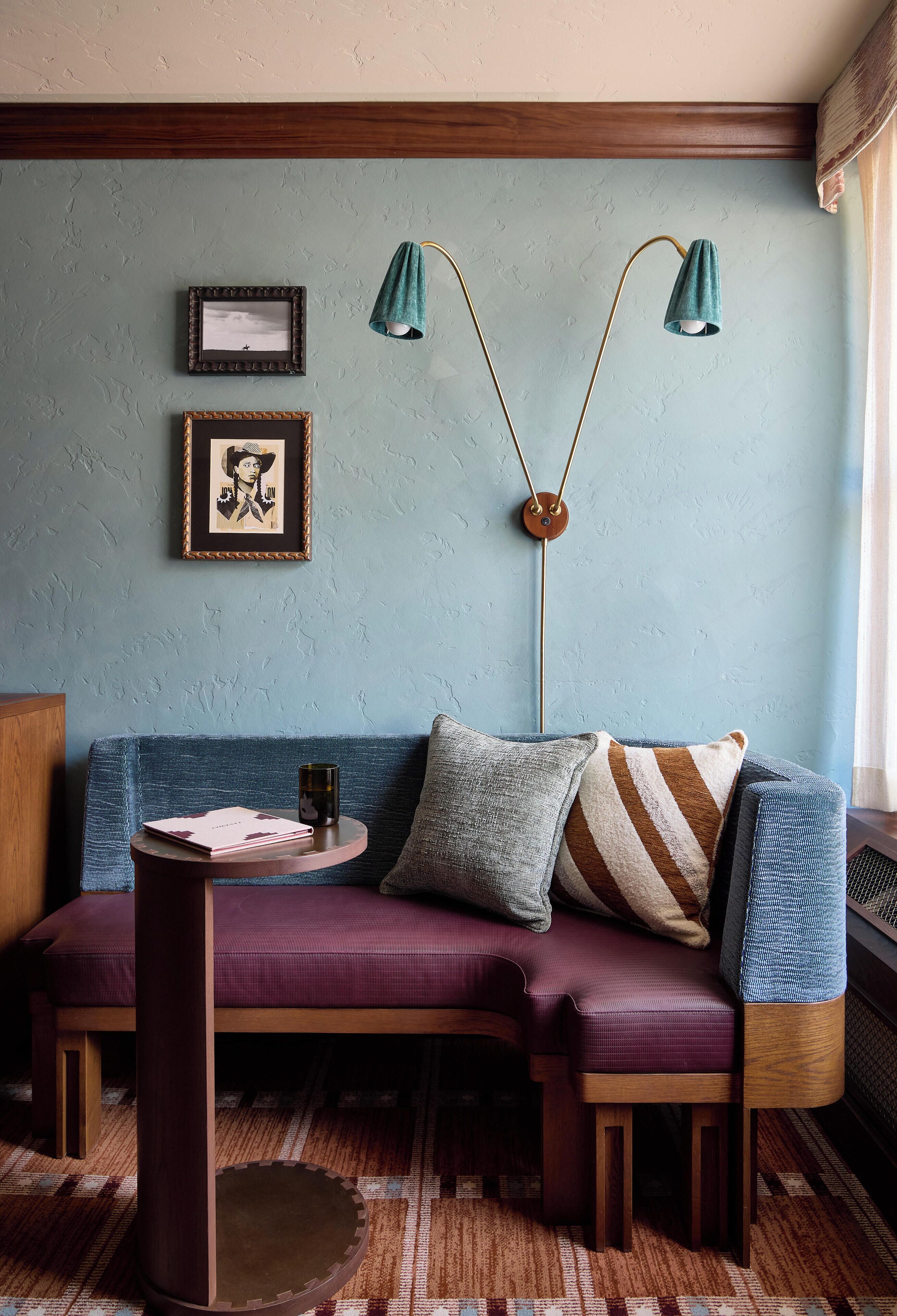 A cozy room with a blue textured wall, a wooden ceiling, a blue sofa with cushions, a small round table with a book and a cup, and a floor lamp with a green shade.