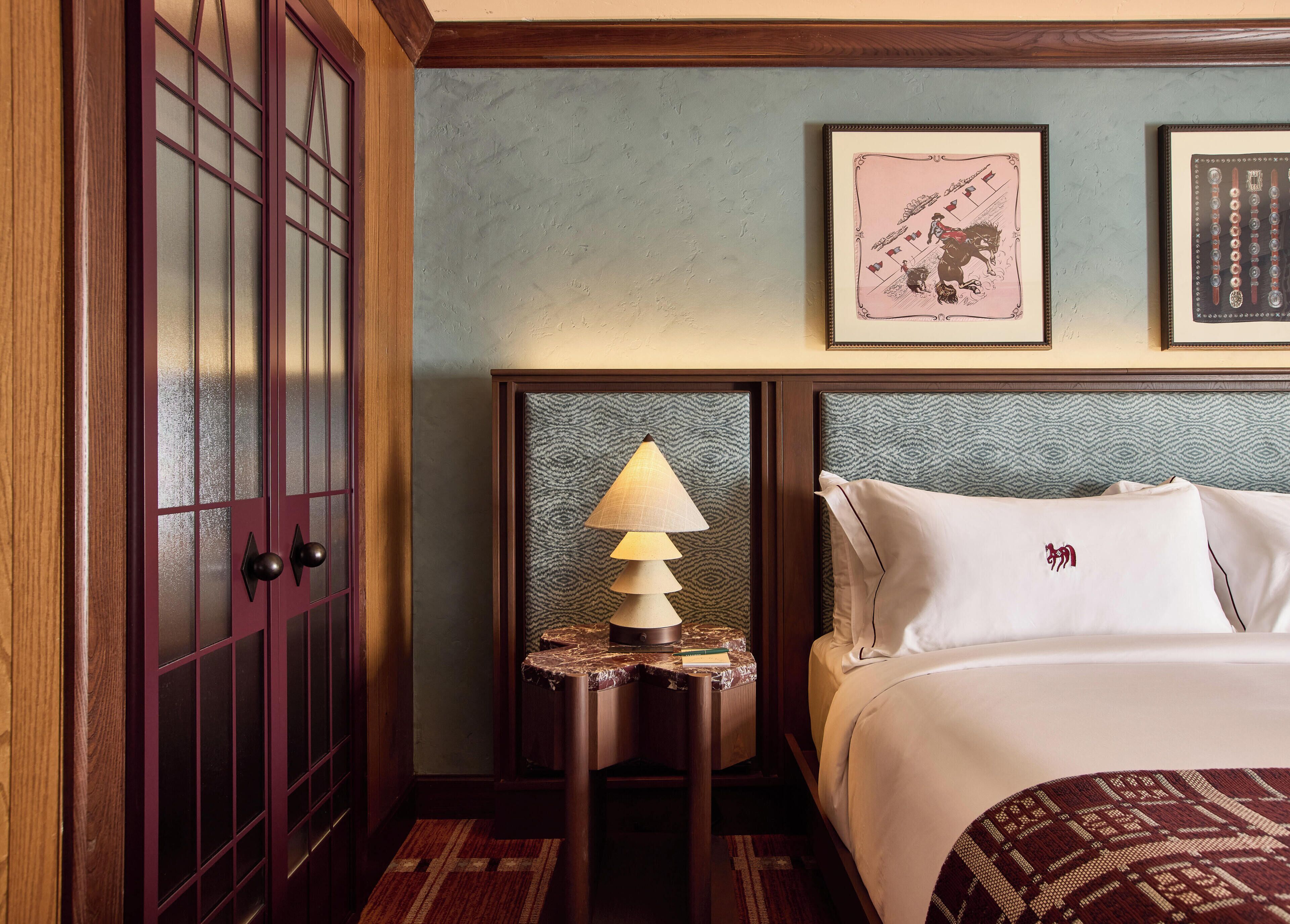 A hotel room with a bed, a nightstand with a lamp, a wooden wardrobe, and two framed pictures on the wall.