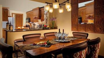 A dining area with a wooden table set for six, surrounded by leather chairs. A tray with glasses and a selection of cheeses and fruits is on the table.
