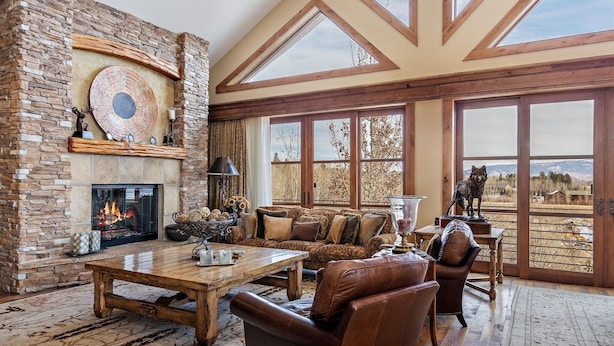 A spacious living room with a stone fireplace, large windows, and a view of the outdoors. - Faraway Jackson Hole (Teton Village)