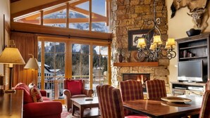 A cozy living room with a stone fireplace, large windows, and a view of mountains. - Faraway Jackson Hole (Teton Village)