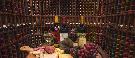 A wine cellar with numerous wine bottles on shelves and a selection of cheeses and grapes on a wooden barrel.