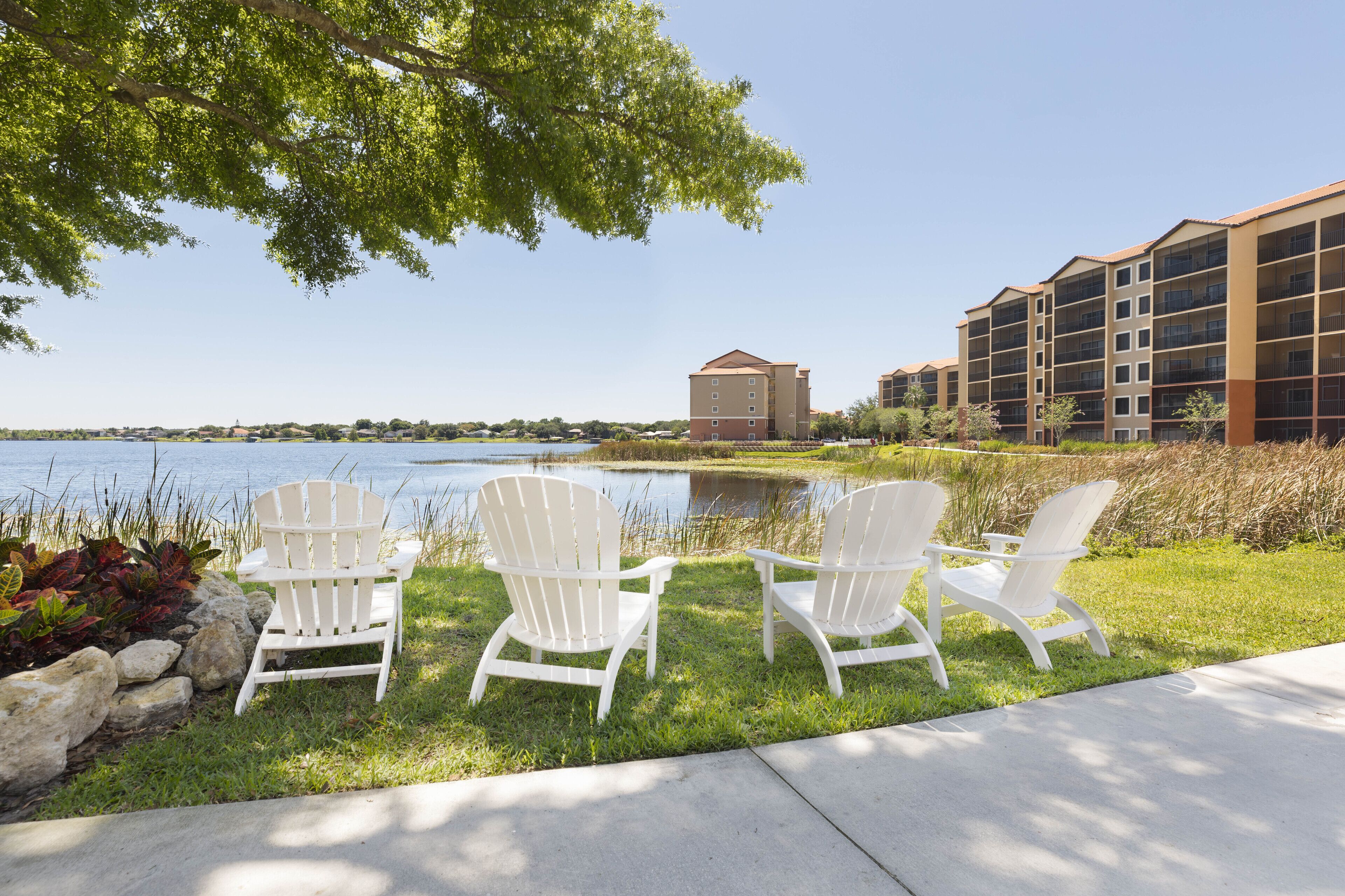 Three white Adirondack chairs by a lake with a building in the background.