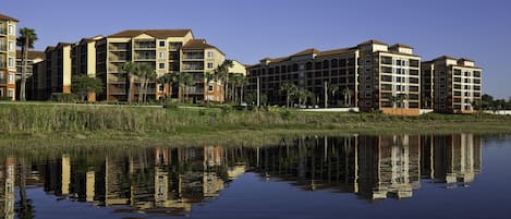 A multi-story hotel with a reflection in the water, surrounded by greenery and palm trees.