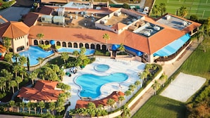 An aerial view of a resort with multiple swimming pools, palm trees, and a large building with a red-tiled roof.