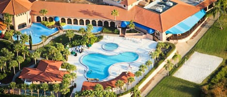 Vista aérea de un complejo turístico con varias piscinas, palmeras y un edificio grande con techo de tejas rojas.