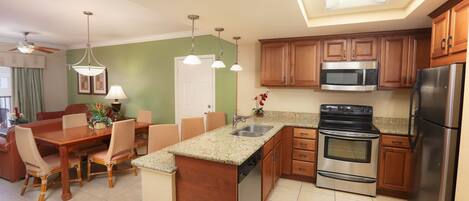 A modern kitchen with wooden cabinets, a stainless steel refrigerator, a microwave, and an oven. There is a dining area with a wooden table and chairs.