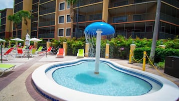 Una piscina con fuente, rodeada de sillones de descanso y un edificio de varios pisos al fondo.
