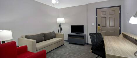 A hotel room with a beige sofa, a red armchair, a wooden desk, a television on a stand, a ceiling fan, and a door with a light switch.