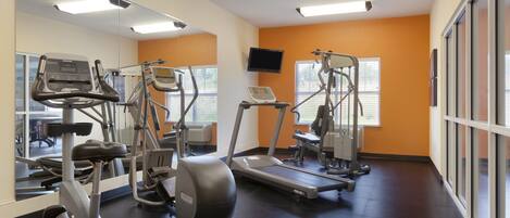 Uma academia com equipamentos de cardio, televisão e janelas amplas.