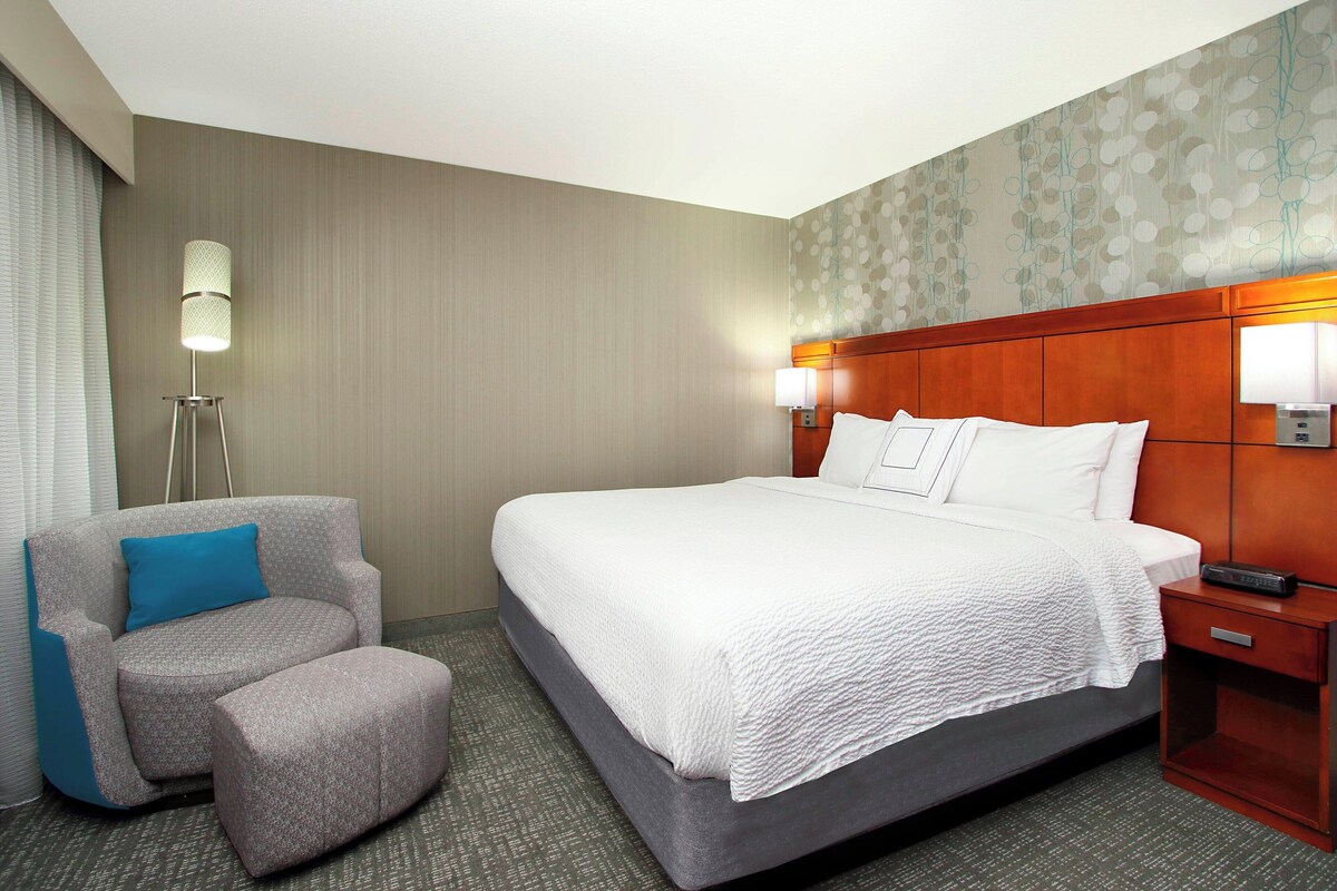 Une chambre d’hôtel avec un grand lit, un fauteuil gris avec un coussin bleu, une table de chevet en bois avec une télécommande, et une lampe fixée au mur.