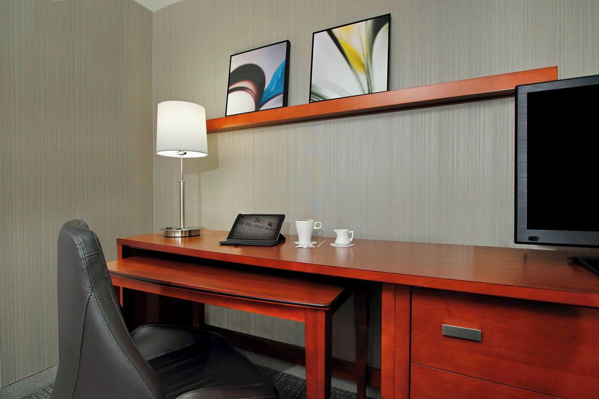 Ein Hotelzimmer mit Holzschreibtisch, einem Stuhl, einer Lampe, einem Flachbildfernseher, einem Laptop und einer Kaffeetasse.