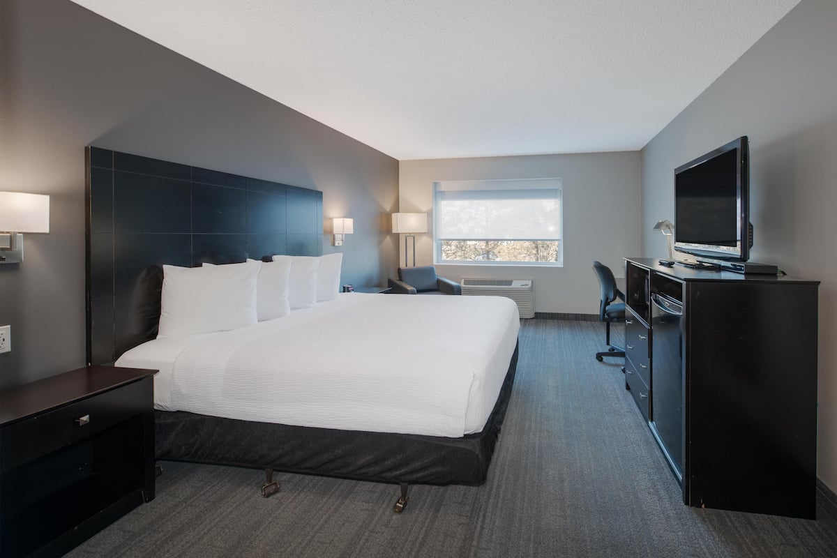 A modern hotel room with a large bed, a desk with a computer, a television, and a window with blinds.